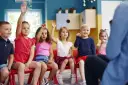 Dzieci na czerwonych krzesłach podnoszą ręce, odpowiadając na pytanie, ile lat ma się w zerówce.