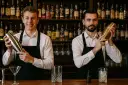 Dwóch barmanów w czarnych fartuchach i białych koszulach trzyma shakery do drinków na tle półek z butelkami alkoholu.
