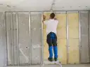Montaż wełny mineralnej na stelażu to kluczowy etap, by jak wyciszyć mieszkanie. Mężczyzna na drabinie dokłada izolację.