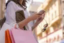 Kobieta z zakupami sprawdza telefon, planując refinansowanie pożyczki.