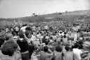 Tysiące ludzi siedzi na trawie, patrząc na scenę festiwalu. To jest Woodstock, gdzie jest muzyka i wolność.
