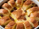 Pudding chlebowy z rodzynkami i cynamonem, pachnący jak domowe wypieki. Obok, w oddali, można dostrzec ślady puddingu ryżowego.