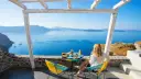 Mujer desayunando en balcón con vista al mar azul y montañas.