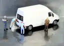 Miniaturowi pracownicy zajmują się białym busem budowlanym. Jeden z nich czyści tył pojazdu, inny trzyma narzędzie, a trzeci obserwuje.