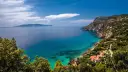 Turquoise water laps a rocky Tuscan coast with villas nestled among trees, under a blue sky with clouds.