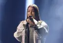 Młoda artystka śpiewa do mikrofonu na scenie. Czy to jej występ na Eurowizji?