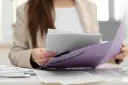 Kobieta w beżowej marynarce przegląda dokumenty w fioletowej teczce, przygotowując się do złożenia wniosku o urlop rodzicielski.