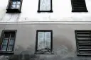 Stara elewacja z widocznymi pęknięciami i zaciekami. Okna z żaluzjami i jedno wybite.