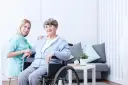 Uśmiechnięta pielęgniarka w turkusowym uniformie wspiera starszą kobietę na wózku inwalidzkim.