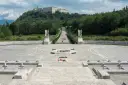 Cmentarz żołnierzy na wzgórzu, z widocznym klasztorem na szczycie. Droga prowadzi do niego przez monumentalny plac z krzyżami i pomnikiem. To miejsce pamięci o bitwie pod **Cassino - trzeci / rzeka**.