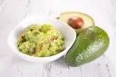 Miseczka z guacamole, obok całe i przekrojone awokado, na białym, drewnianym tle.