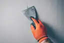 Ręka w pomarańczowej rękawicy nakłada masę na pęknięcia ścian za pomocą szpachelki.