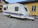 Biała przyczepa kempingowa Hobby 460 UFE "de luxe easy" stoi na kostce brukowej przed żółtym budynkiem.