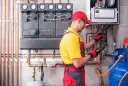 Instalator wykonuje próbę szczelności instalacji gazowej powietrzem, używając detektora. W tle widoczny kocioł i manometry.