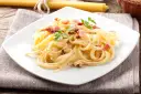 Pyszne spaghetti carbonara z boczkiem i natką pietruszki na białym talerzu.