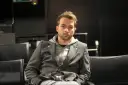 Marcin Hycnar w szarej marynarce i bluzie z kapturem siedzi na czarnym krześle, wpatrując się wprost przed siebie.