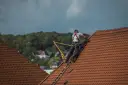 Dekarz w szelkach asekuracyjnych pracuje na czerwonym dachu.