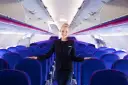 Uśmiechnięta stewardessa Wizz Air stoi w rzędach niebieskich foteli w samolocie.