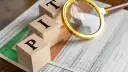 Drewniane klocki z literami "P", "I", "T" leżą na dokumentach podatkowych. Lupa sugeruje analizę, by osiągnąć cel: rozliczenie wolne od podatku.