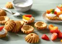 Kruche babeczki, niektóre puste, inne z kremem i owocami, na zielonym tle.
