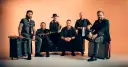 Sechs Männer einer Band posieren vor orangefarbenem Hintergrund, einige sitzen, einer steht.