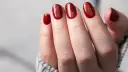 Glitzernde rote Gel-Nagel-Designs auf einer Hand, die aus einem grauen Pullover ragt.