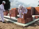 Dwóch budowlańców w białych kombinezonach układa cegły na fundamencie.