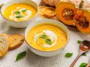 Aksamitna zupa z dyni hokkaido, udekorowana śmietaną i bazylią, podana z grzankami. Obok połówka dyni.