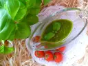Aromatyczne pesto bazyliowe w szklanej sosjerce, ozdobione listkami bazylii, obok gałązki pomidorków koktajlowych.