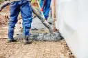 Robotnicy w niebieskich ogrodniczkach i gumowcach wylewają beton. Widać, ile piasku na kubik betonu jest potrzebne do budowy.