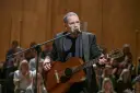 Artysta w okularach gra na gitarze podczas koncertu Męskie Granie. Publiczność w tle.