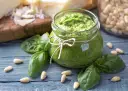 Słoik domowego pesto z bazylii, otoczony świeżymi liśćmi bazylii i orzeszkami pinii. Obok kawałki parmezanu. Idealne do makaronu.