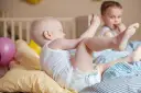 Dzieci bawią się na łóżku. Maluch w pieluszce sięga po zabawkę, a starszy chłopiec je. Ciekawe, ile pieluch miesięcznie zużywają takie energiczne maluchy!
