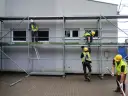 Monterzy w kaskach i kamizelkach pracują na rusztowaniach przy budynku.