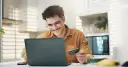 Uśmiechnięty mężczyzna z kartą płatniczą przed laptopem, być może sprawdza limit w koncie przed zakupami online.