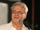 Andrzej Błaszczak, aktor, w okularach i białej koszuli, przemawia do mikrofonu.