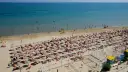A crowded beach with rows of umbrellas and sunbathers, and people swimming in the blue sea.