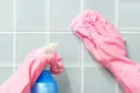 Ręce w różowych rękawiczkach czyszczą niebieskie kafelki sprayem i ściereczką.