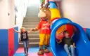 Mascot and two children in a colorful indoor playground with a slide and stairs.