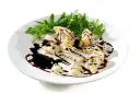 Kawałki parmezanu polane gęstą glazurą balsamiczną, podane z rukolą na białym talerzu.