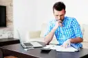 Mężczyzna analizuje dokumenty, zastanawiając się nad prolongatą pożyczki. Na stole laptop, telefon i kalkulator.
