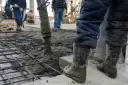 Betoniarka wylewa beton na zbrojenie, pracownicy w gumowcach nadzorują proces.