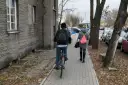 Osoba na rowerze i kobieta idąca chodnikiem obok budynku i drzew.