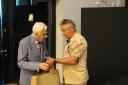 Zdzisław Kuźniar syn, starszy pan w niebieskiej marynarce, wręcza prezent młodszemu mężczyźnie w beżowej koszulce.