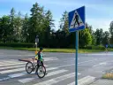 Rowerzystka w kasku prowadzi rower po pasach dla pieszych obok znaku drogowego.