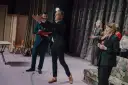 Arkadiusz Brykalski w garniturze gestykuluje na scenie, obok niego mężczyzna z teczką. W tle kobiety w eleganckich strojach.