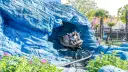 Personas en un coche de montaña rusa saliendo de una cueva azul rocosa, con palmeras y un paisaje de cuento de hadas al fondo.