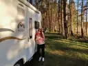 Kobieta z kubkiem kawy opiera się o kampera na leśnej polanie. Wspaniała przygoda w **Finlandii kamperem**!