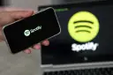 Dłoń trzyma smartfon z logo Spotify. W tle ekran laptopa z tym samym logo.
