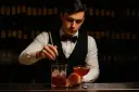 Barman w eleganckim stroju miesza drinka z lodem w szklanicy, obok stoi gotowy koktajl.